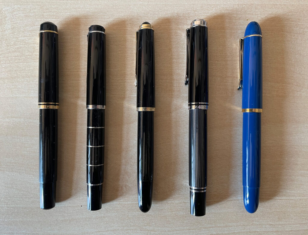 Picture of five Pelikan fountain pens, four black ones and one blue one.
